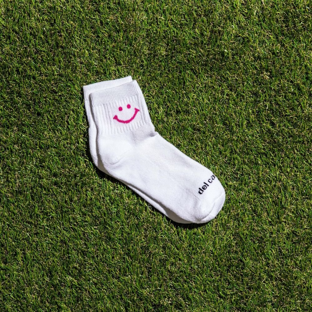 White Del Campo Golf sock with pink smiley face design on grass. | on-course, product