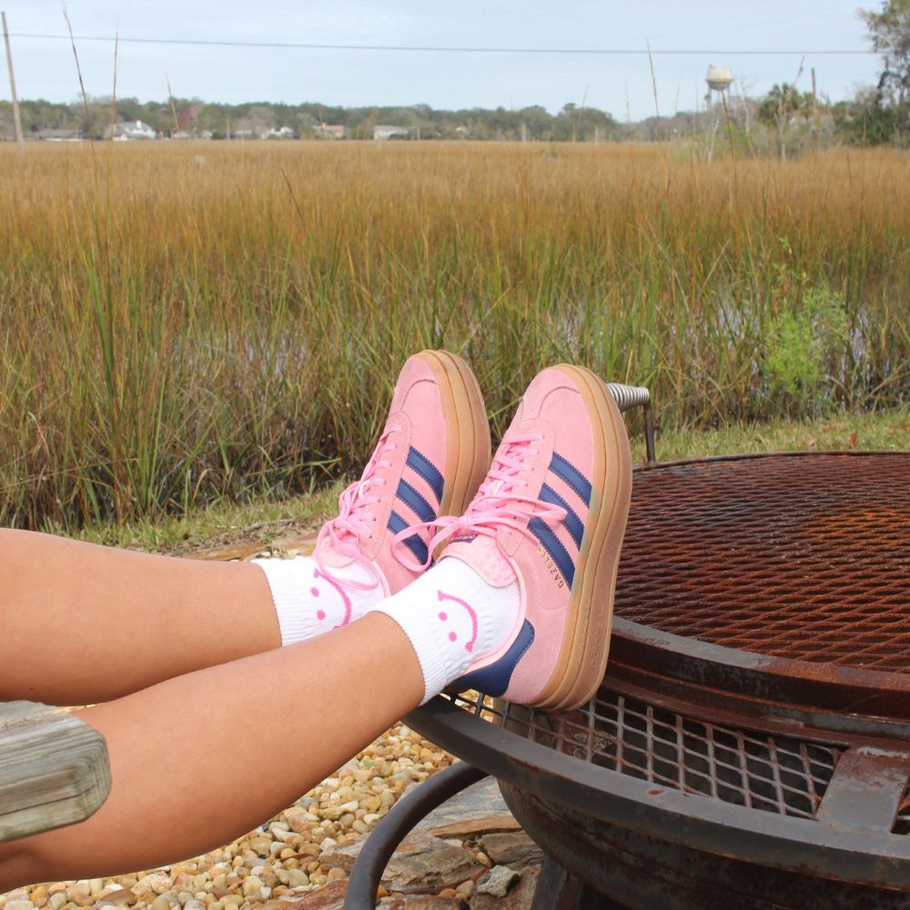 Person wearing white Del Campo Golf socks with pink smiley design and pink sneakers, relaxing by a waterfront with marshland in the background. | women, lifestyle