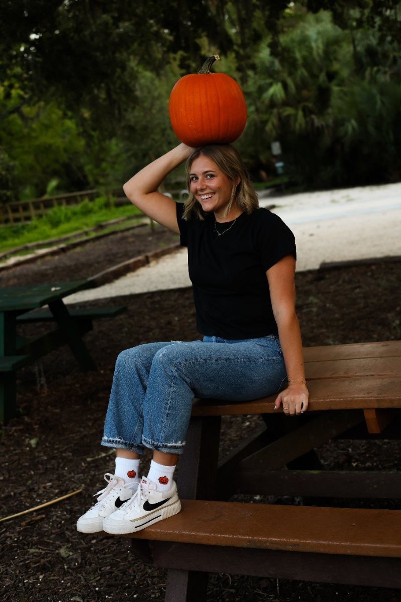 Smiling woman balancing a pumpkin on her head, wearing white Del Campo socks with a jack-o'-lantern design