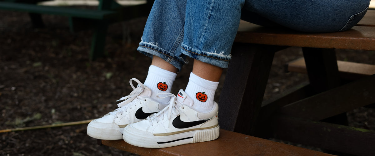 White Del Campo socks with a jack-o'-lantern design, worn with white sneakers on a picnic bench