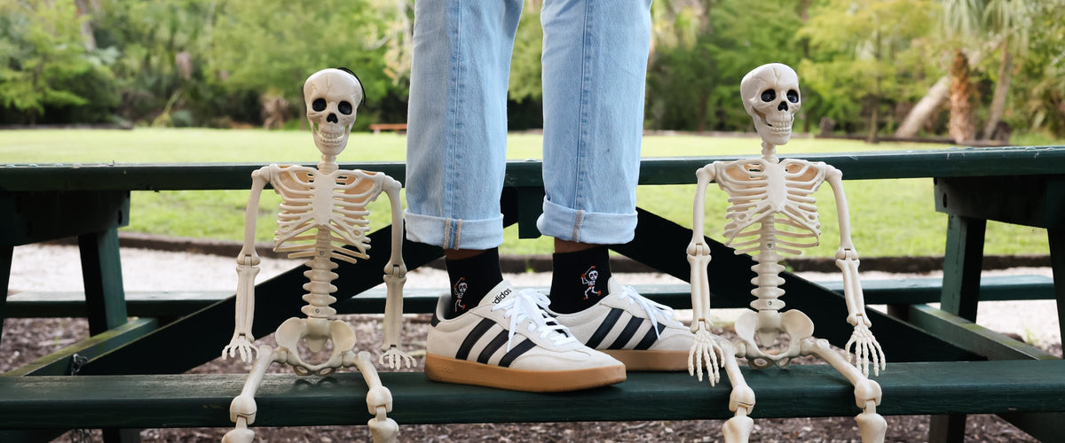 Black Del Campo socks with a skeleton design, standing between two toy skeletons on a picnic table