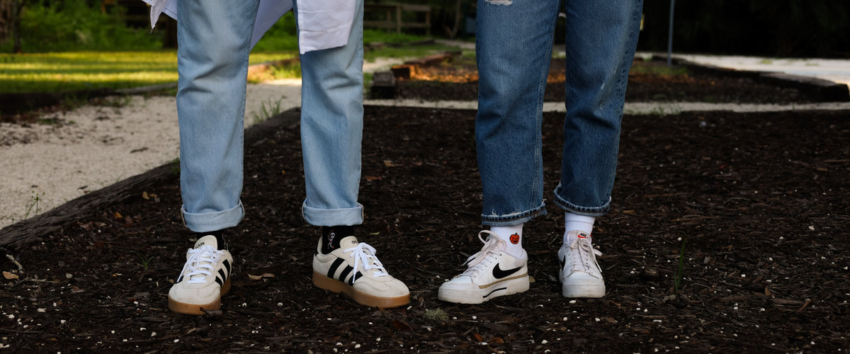 Two people standing on mulch, one in black Del Campo Halloween socks, one in white Del Campo socks with a jack-o'-lantern design