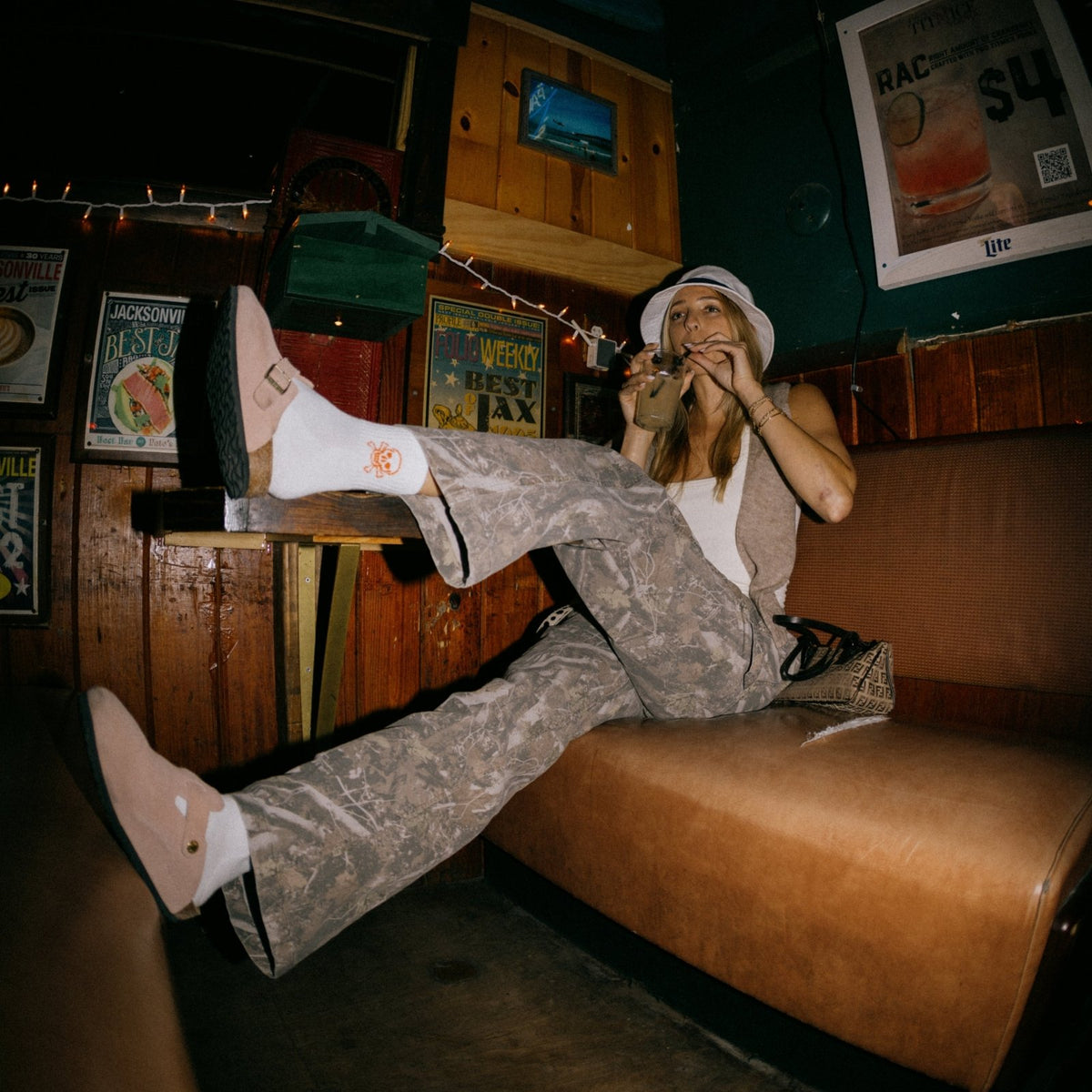 Woman wearing white Del Campo Golf Crossbones socks in a casual bar setting, feet up on a leather ottoman. | women, lifestyle