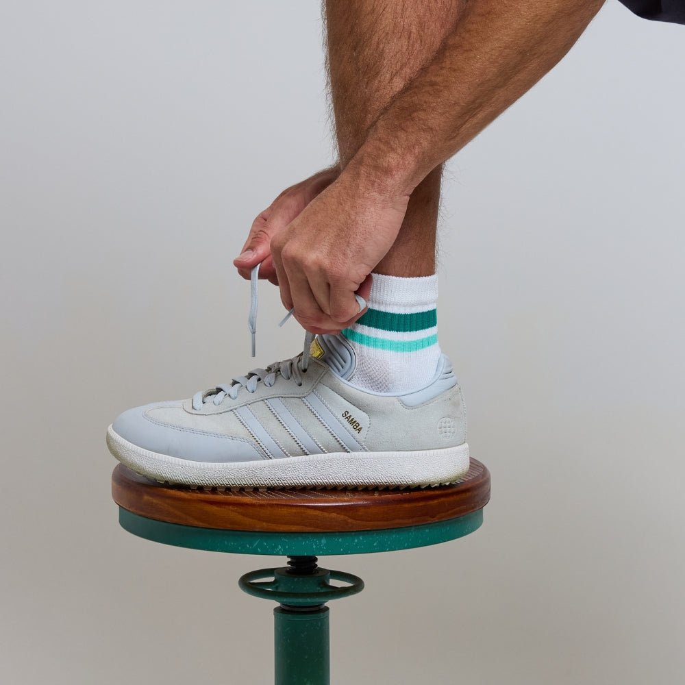 Man wearing white Del Campo Golf socks with green and turquoise stripes, standing on a golf course tee marker. | men, on-course, lifestyle
