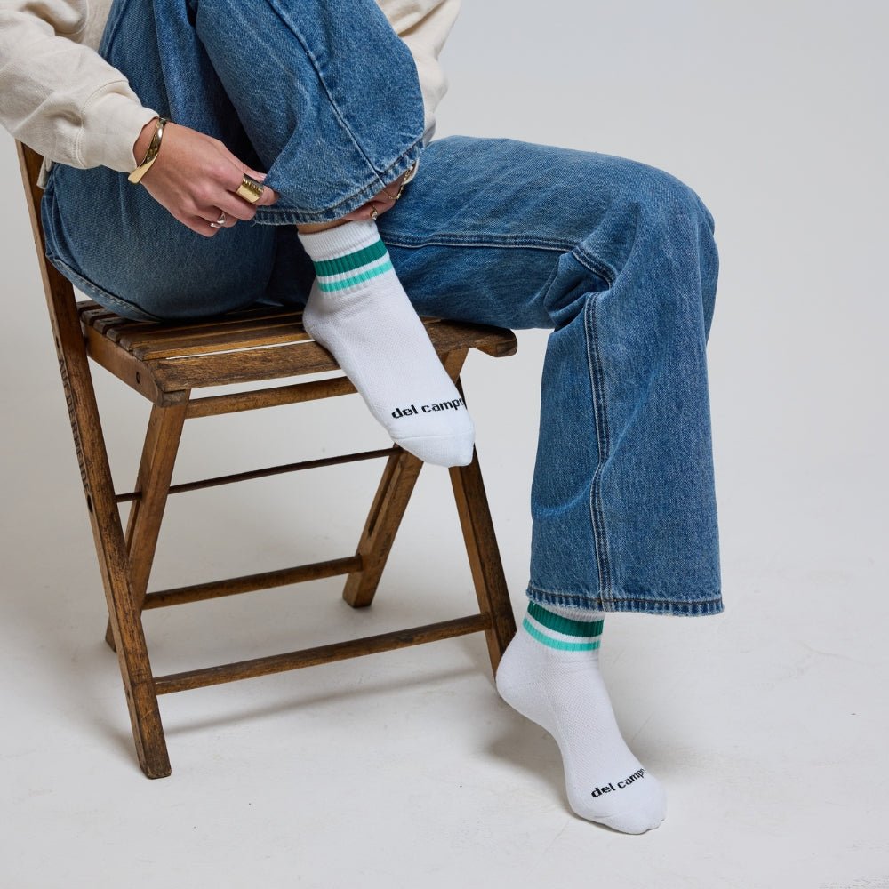 Person wearing white Del Campo Golf socks with green and turquoise stripes, sitting on a wooden chair in jeans. | lifestyle, women