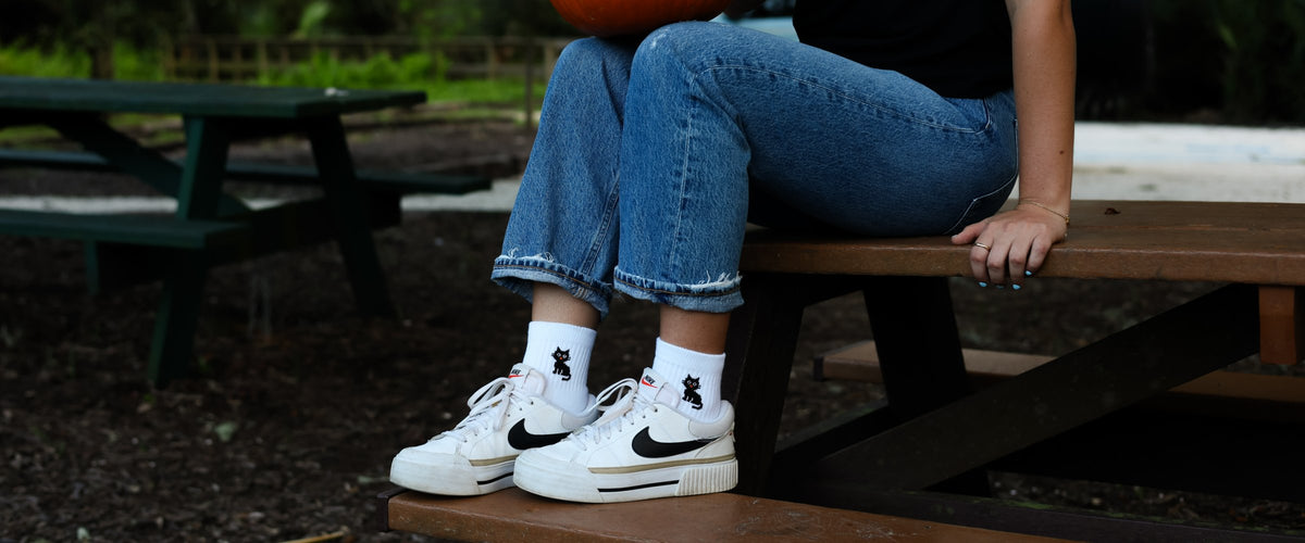 White Del Campo socks with a black cat design, worn by a woman holding a pumpkin on a picnic bench