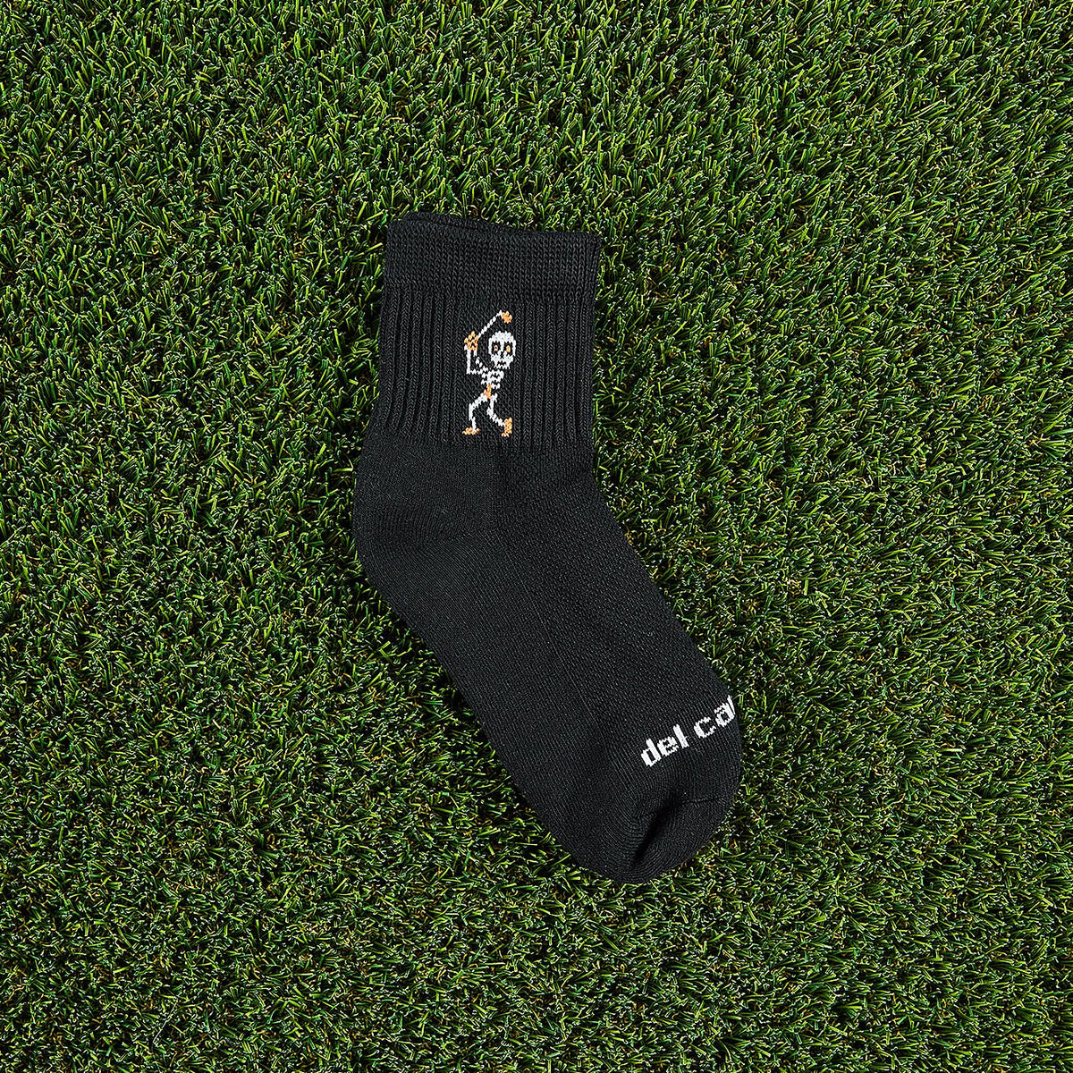 Del Campo Swingin' Skelly Halloween golf socks, black quarter socks with a skeleton golfer mid-swing in white and orange, single sock on grass